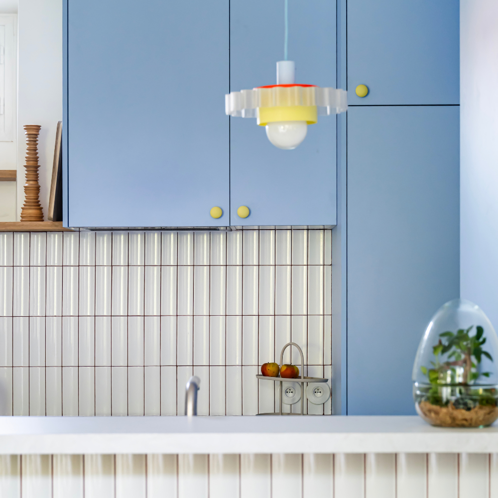 Cuisine Ikea Metod aux portes bleu clair coloris "Piscine" Studio Couleur, et boutons de porte jaune mat. Crédence en céramique et plan de travail blanc.