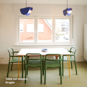 Table de salle à manger rose clair créée sur-mesure avec des panneaux Studio Couleur. Pieds de table jaune-orangé et chaises vertes. Suspensions bleu électrique.